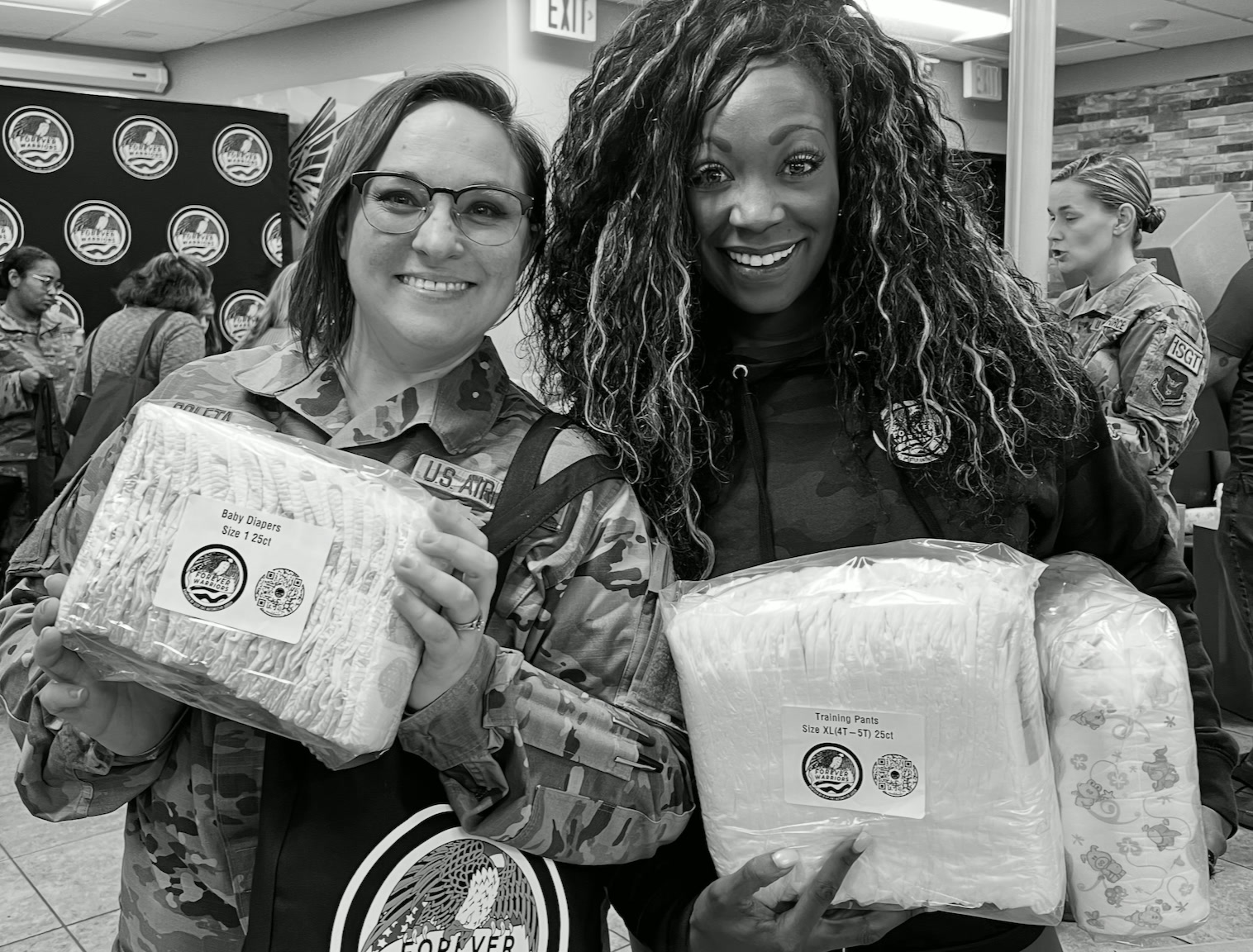 Two women holding packaged baby diapers and training pants at a military diaper distribution event, featured in a guide to whether WIC covers diapers.