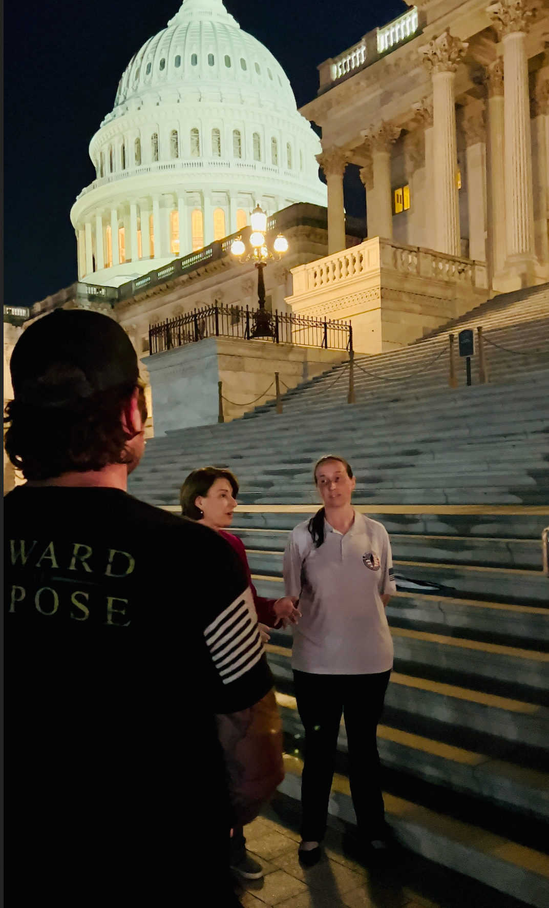 People speaking on the U.S. Capitol steps at night, the lit Capitol dome and columns behind them.