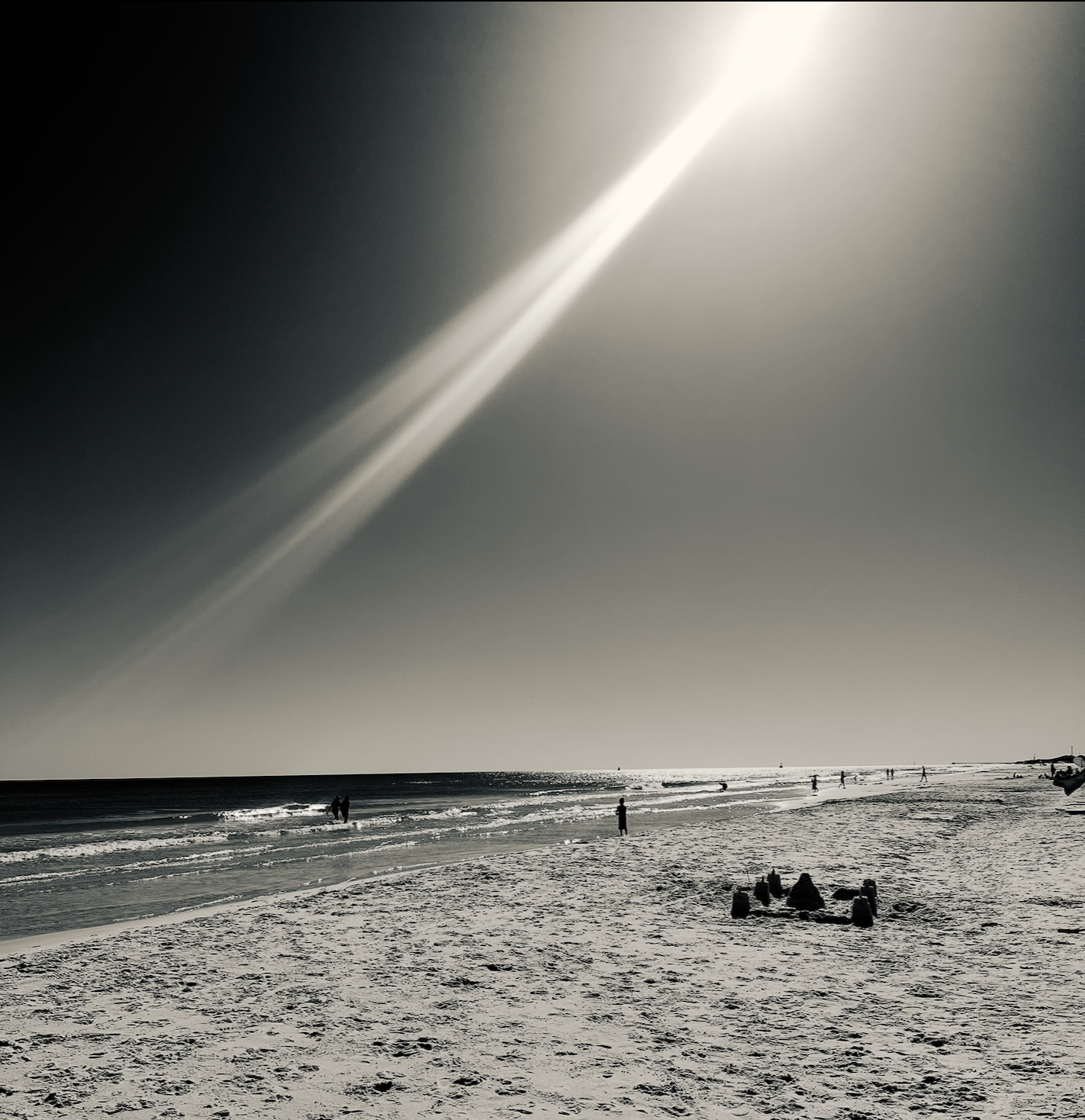 A black-and-white beach on Florida's Emerald Coast, a shaft of sunlight over the ocean, with a few people and a sandcastle on the sand.