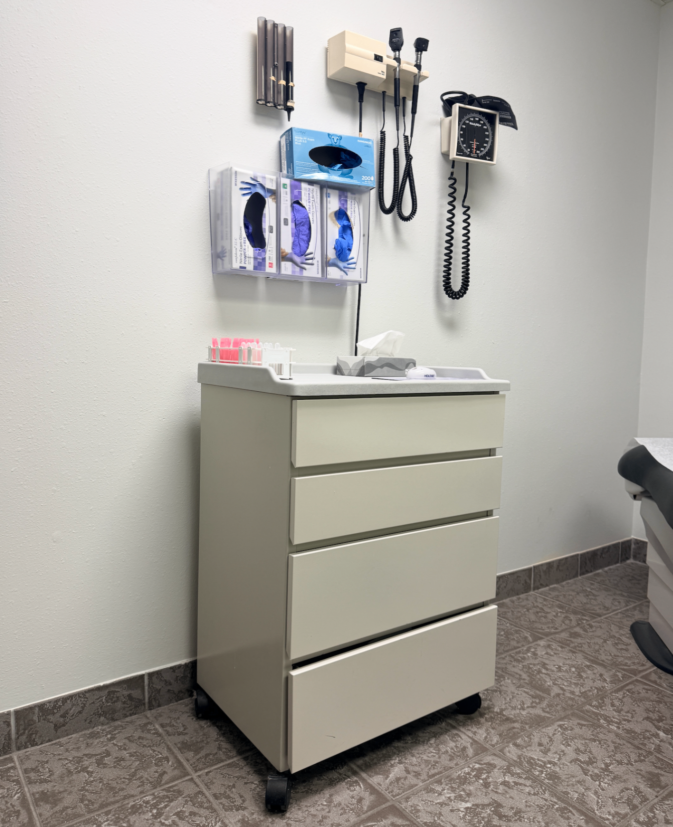 A clean medical exam room with wall-mounted diagnostic instruments and a supply cabinet.